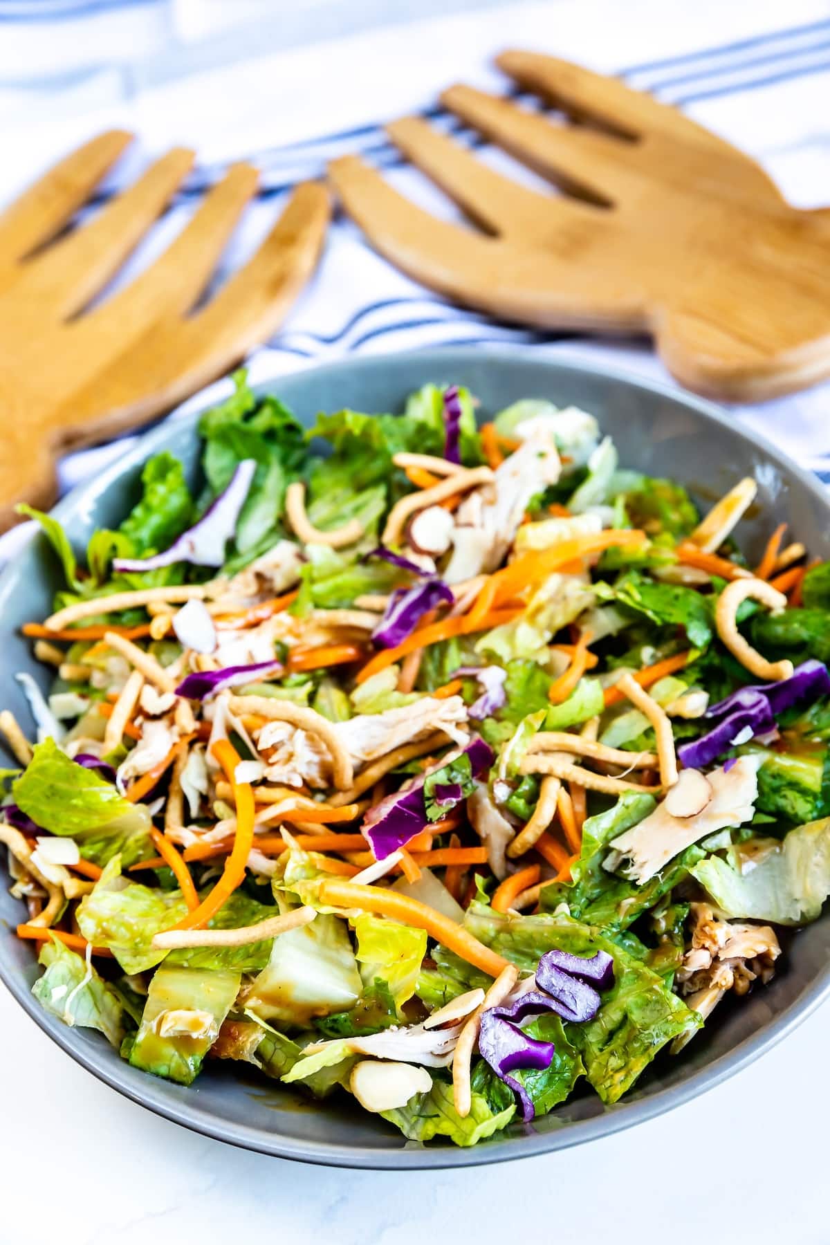 A bowl of Asian-inspired salad with romaine lettuce, shredded carrots, purple cabbage, sliced almonds, chow mein noodles, and shredded chicken, with wooden salad tongs and a striped towel in the background.