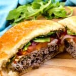 A close-up of a hamburger cut in half, showing layers of ground beef, melted cheese, lettuce, tomato, and red onion on a wooden surface, with fresh arugula and a blue cloth in the background.