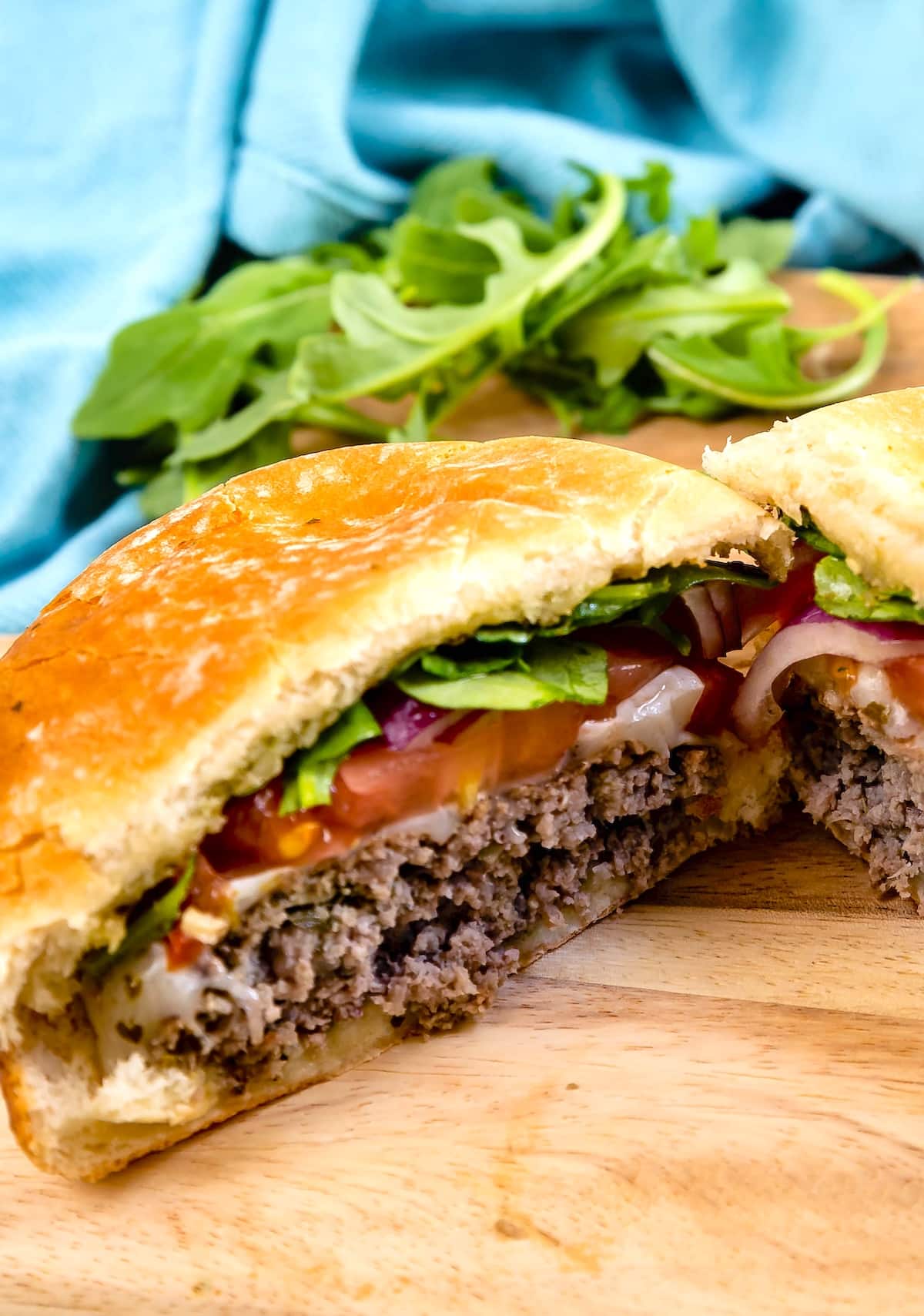 A close-up of a hamburger cut in half, showing layers of ground beef, melted cheese, lettuce, tomato, and red onion on a wooden surface, with fresh arugula and a blue cloth in the background.
