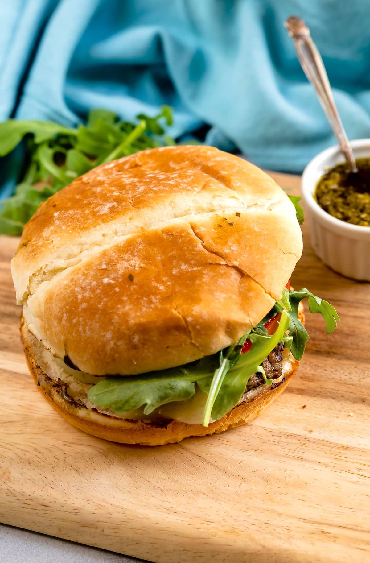 A sandwich with leafy greens and cheese on a round bun sits on a wooden board. There is a small bowl of sauce and a blue cloth in the background.