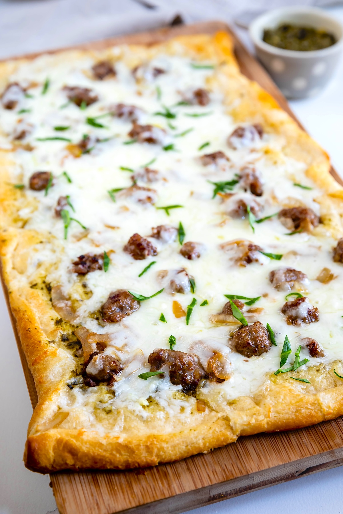 A rectangular pizza topped with melted cheese, sausage pieces, herbs, and a light sauce, served on a wooden board with a small bowl of pesto in the background.