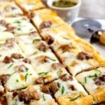 Rectangular slices of cheesy flatbread topped with sausage and herbs are arranged on a wooden board. A pizza cutter and a small bowl of green dipping sauce are in the background.
