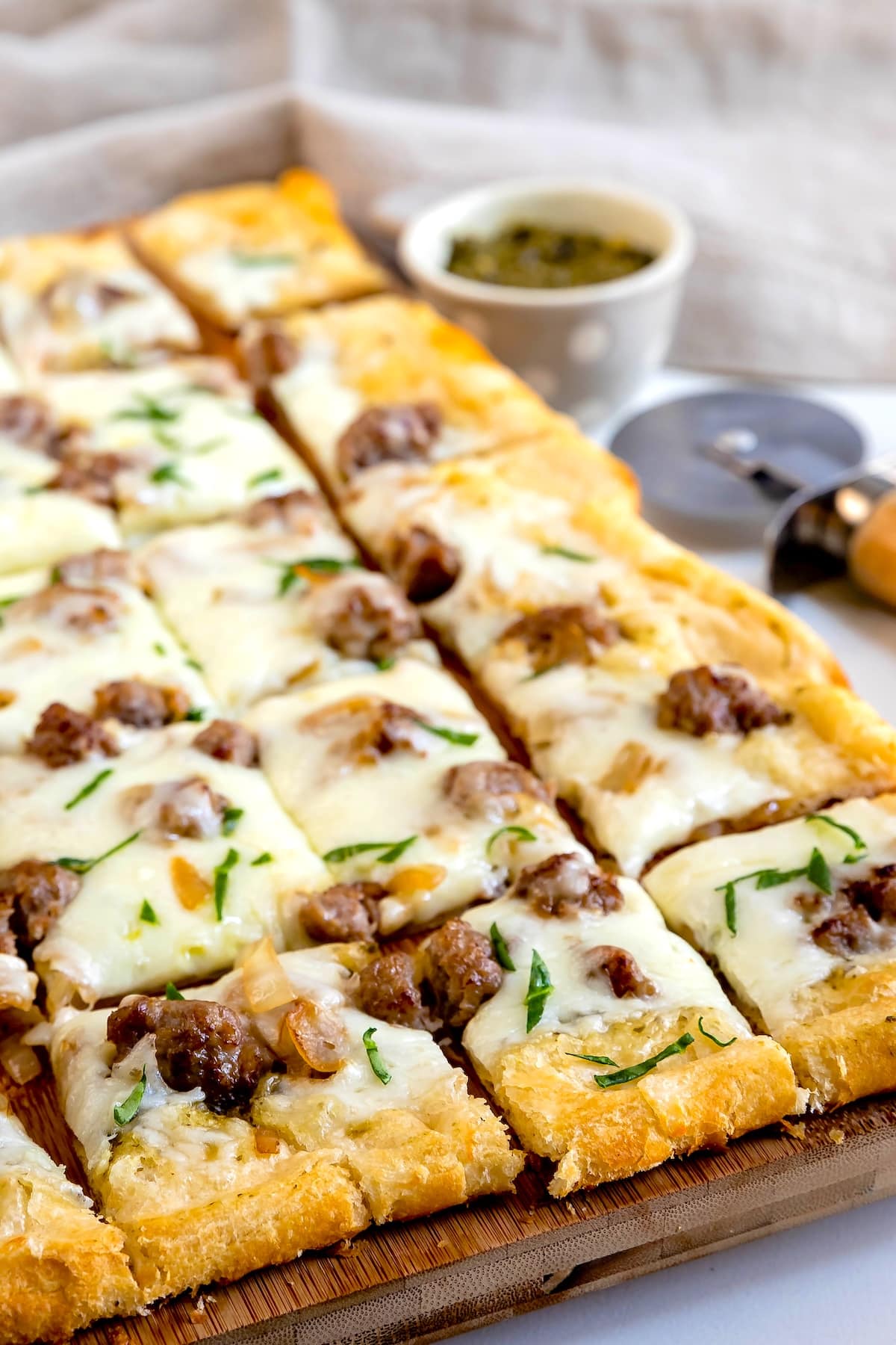 Rectangular slices of cheesy flatbread topped with sausage and herbs are arranged on a wooden board. A pizza cutter and a small bowl of green dipping sauce are in the background.