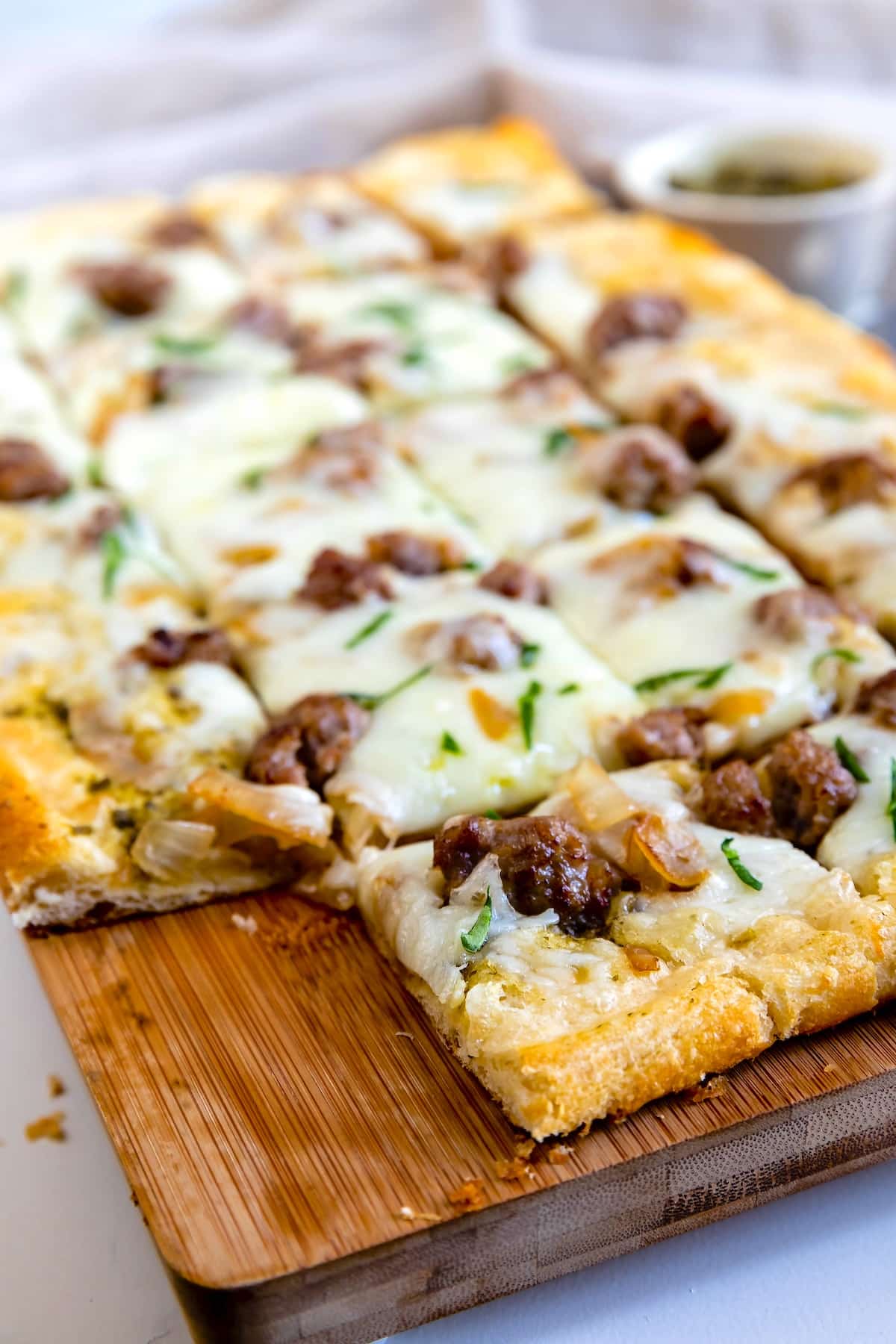 Rectangular flatbread pizza on a wooden board, topped with melted cheese, sausage pieces, and chopped herbs, sliced into squares and ready to serve.
