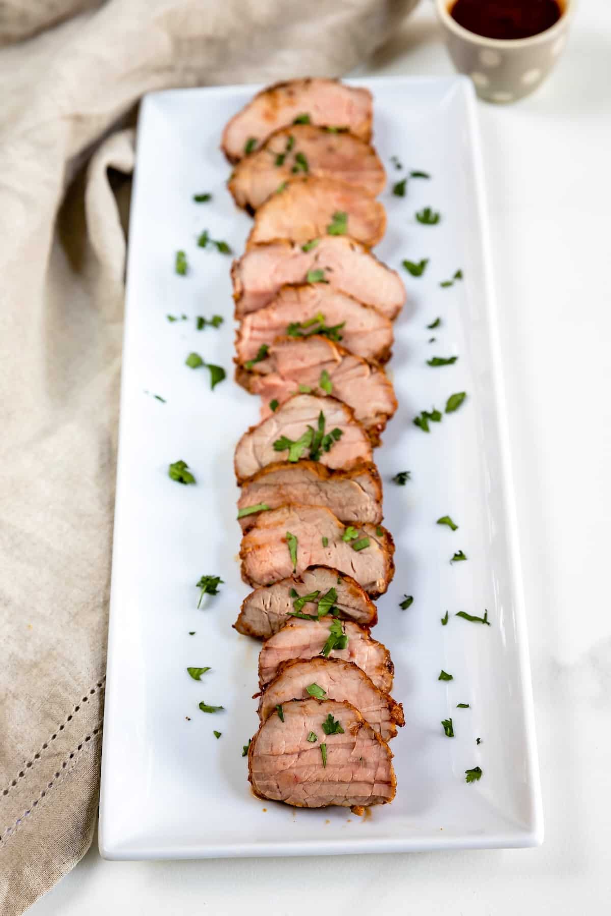 Sliced pork tenderloin arranged neatly on a rectangular white platter, garnished with chopped herbs, with a beige napkin beside the plate.