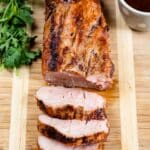 Grilled pork tenderloin sliced on a wooden cutting board, with three pieces cut, fresh parsley on the side, and a cup of sauce in the background.