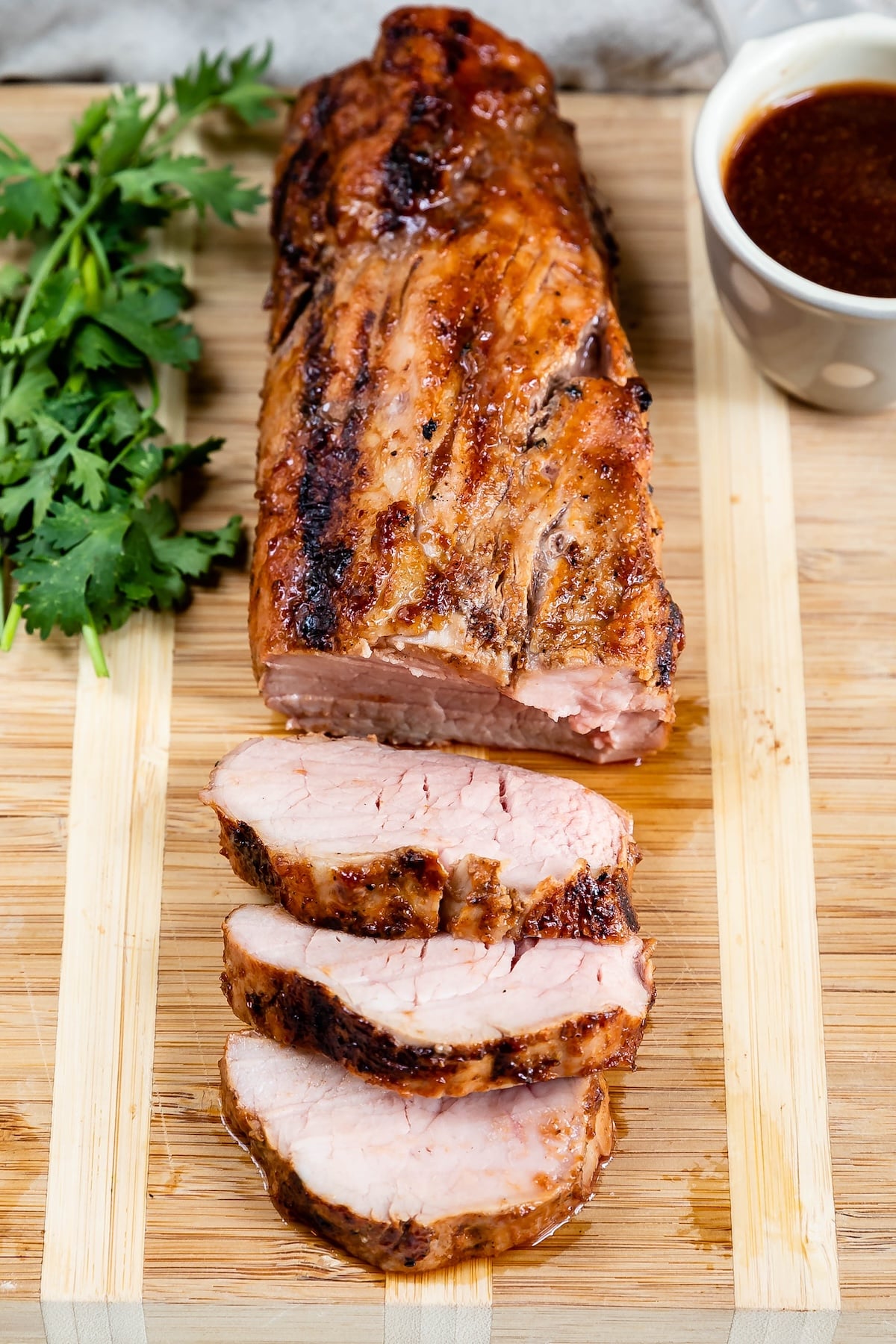 Grilled pork tenderloin sliced on a wooden cutting board, with three pieces cut, fresh parsley on the side, and a cup of sauce in the background.