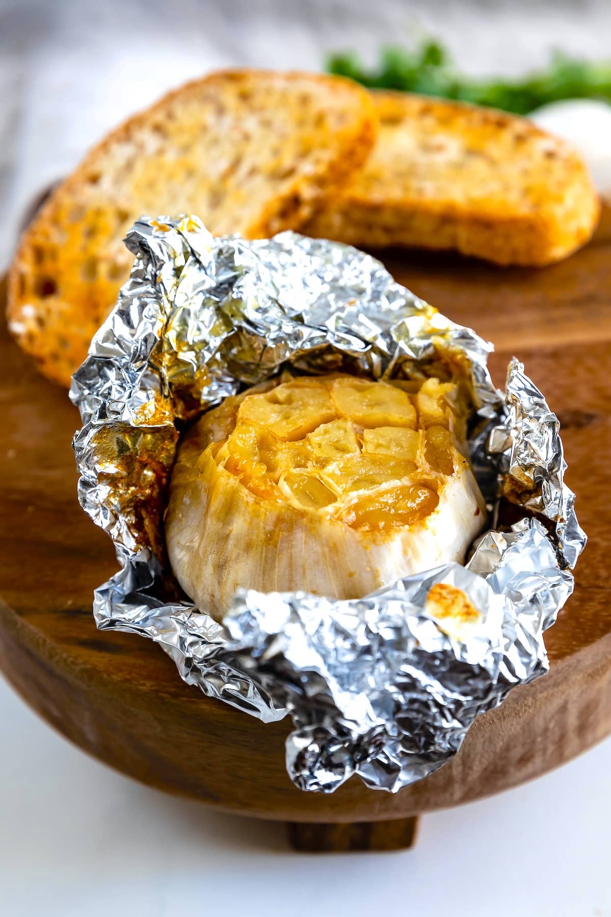 A roasted garlic bulb wrapped in aluminum foil sits on a wooden board, with toasted slices of bread in the background.