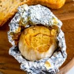 A head of roasted garlic partially wrapped in crinkled aluminum foil sits on a wooden board next to two slices of toasted bread.