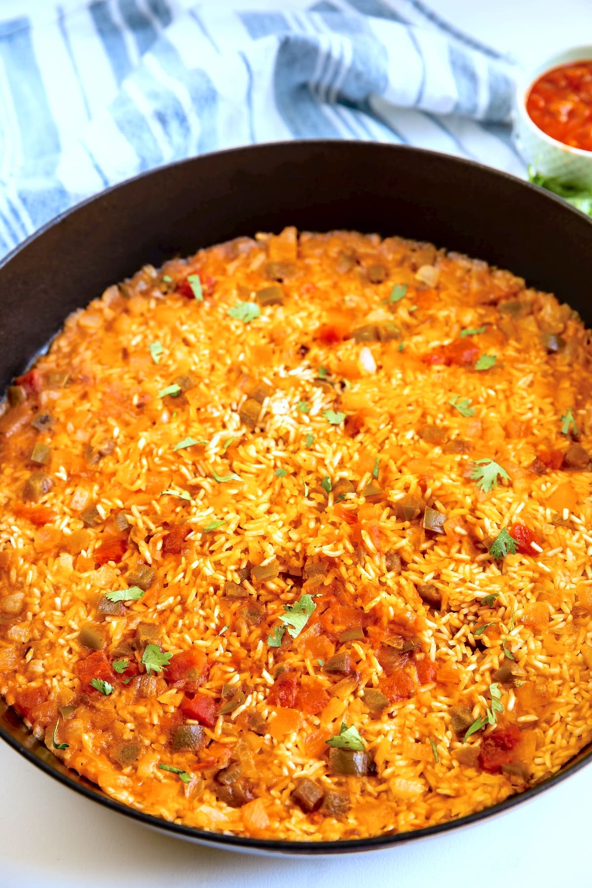 A black skillet filled with baked Spanish rice, garnished with chopped herbs, sits on a white surface with a folded blue-and-white striped towel and a small bowl of sauce in the background.