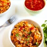 A bowl of rice mixed with vegetables and garnished with chopped herbs sits on a white surface. Nearby are a fork, fresh cilantro, a bowl of tomato-based sauce, and another bowl of the same rice dish.