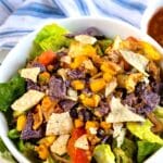 A bowl of salad topped with lettuce, tomatoes, corn, black beans, shredded cheese, and crushed blue and white tortilla chips, with a striped cloth and a small bowl of salsa in the background.