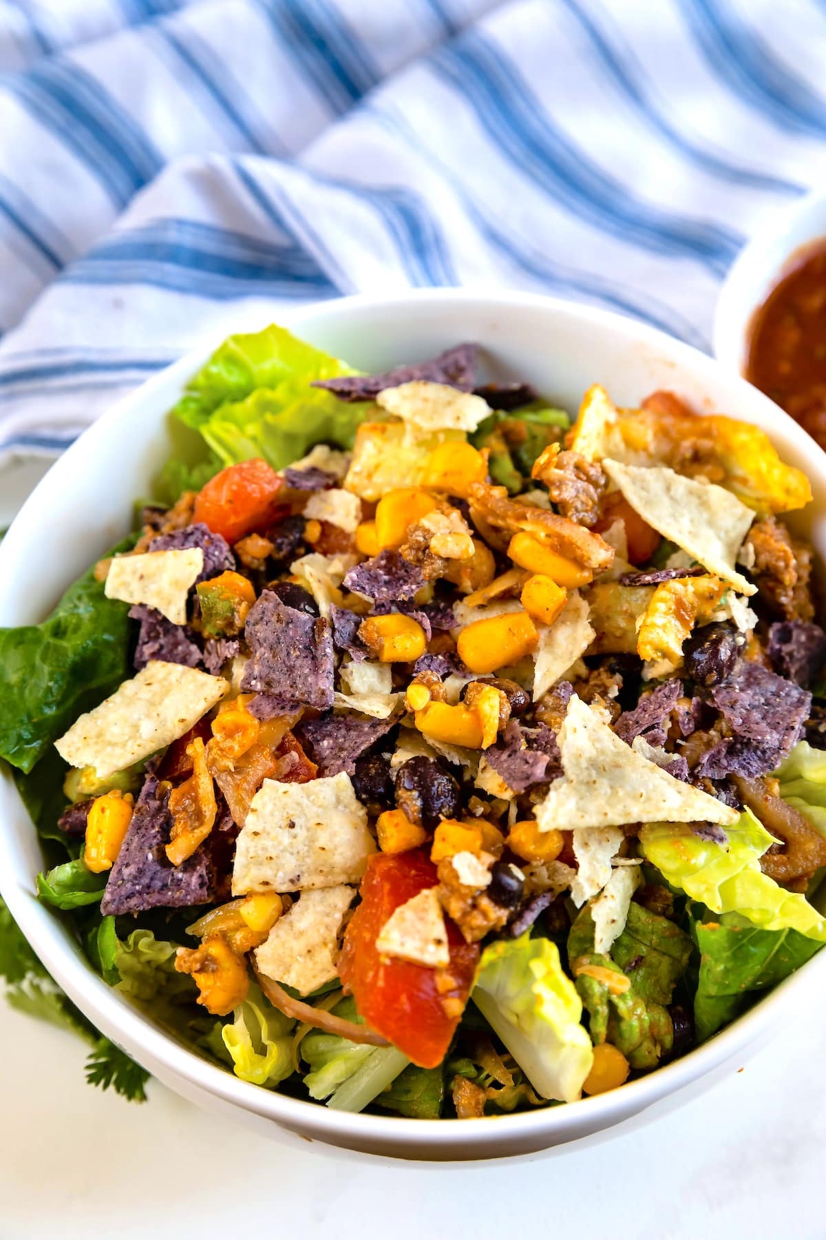 A bowl of salad topped with lettuce, tomatoes, corn, black beans, shredded cheese, and crushed blue and white tortilla chips, with a striped cloth and a small bowl of salsa in the background.