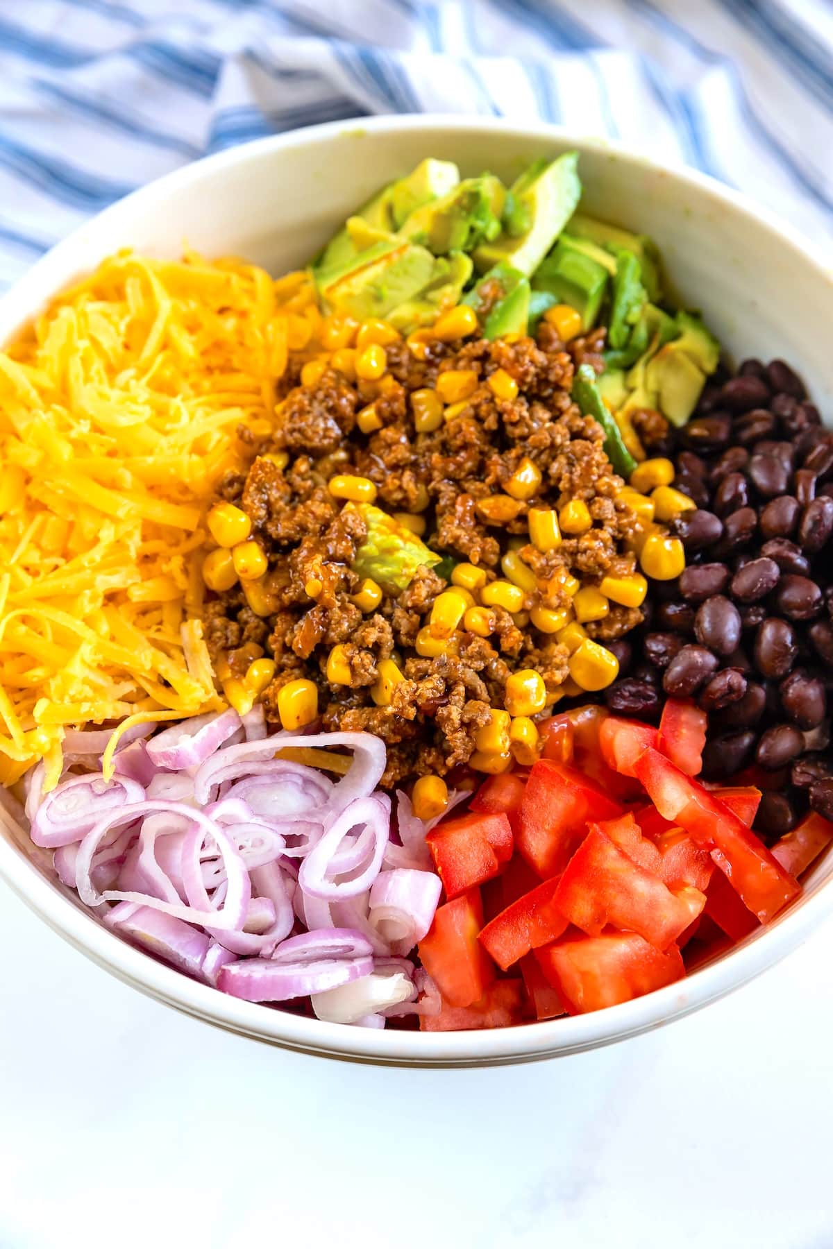 A bowl filled with shredded cheese, sliced avocado, black beans, seasoned ground beef with corn, sliced red onions, and diced tomatoes arranged in sections. A striped cloth is in the background.