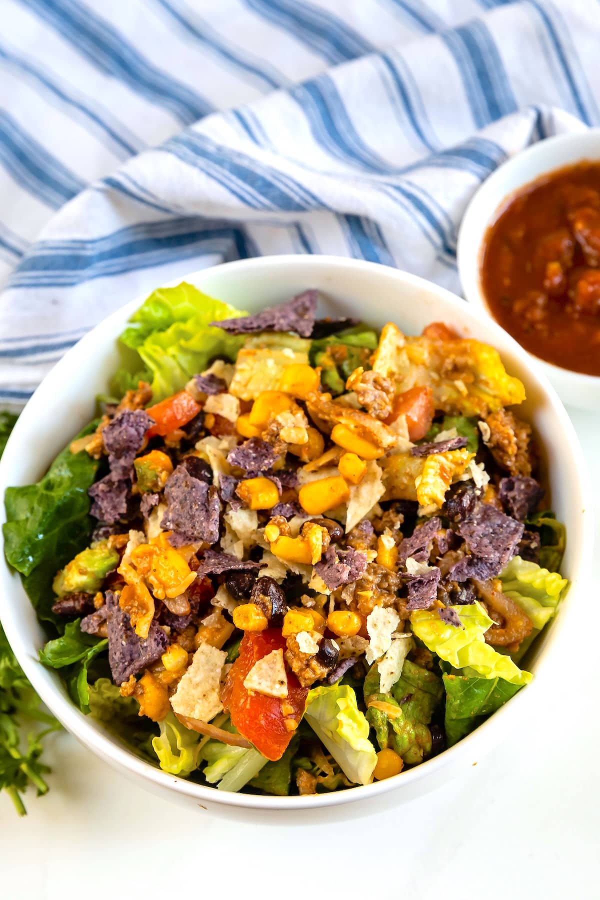 A white bowl filled with a colorful salad of romaine lettuce, tomatoes, corn, crushed blue and yellow tortilla chips, and cheese. A small bowl of salsa and a striped cloth are in the background.