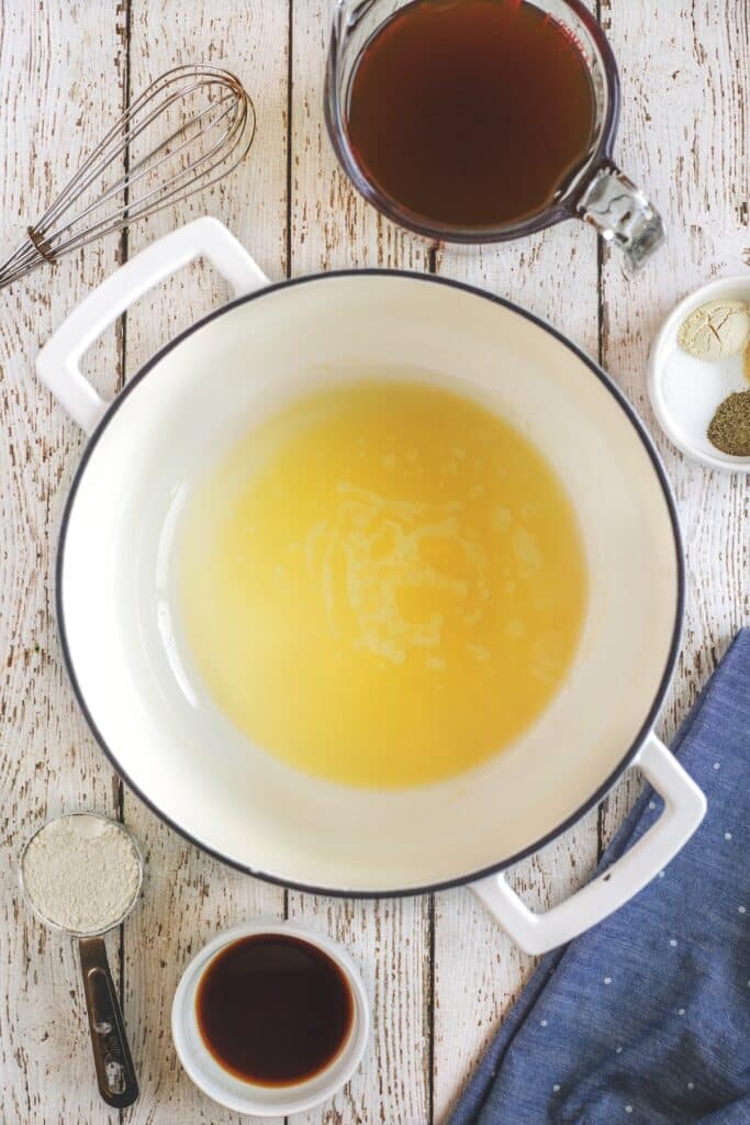 A white pot with melted butter sits on a wooden surface surrounded by a whisk, a glass measuring cup of broth, small bowls of spices, a measuring cup of flour, and a small bowl of soy sauce.