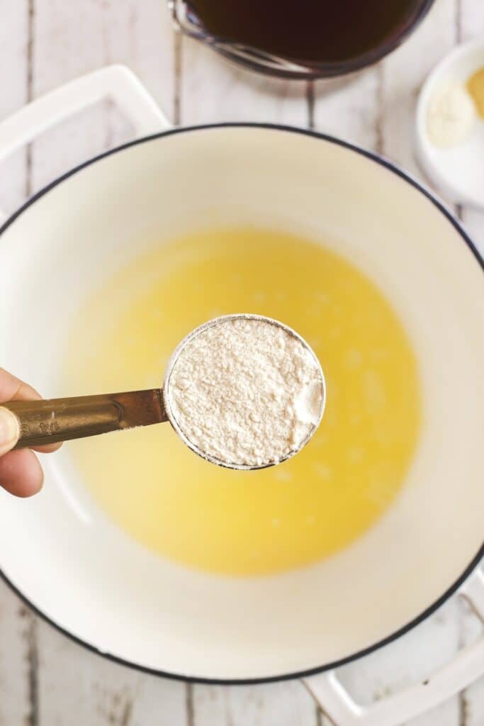 A hand holds a measuring cup of flour above a white pot filled with melted butter, ready to be mixed; other ingredients are blurred in the background on a wooden surface.