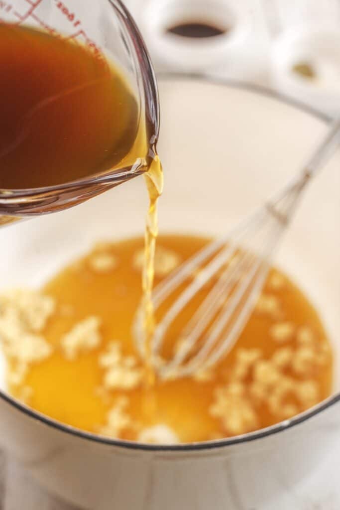 A close-up of broth being poured from a measuring cup into a pot with minced garlic and a whisk, preparing to cook a dish.