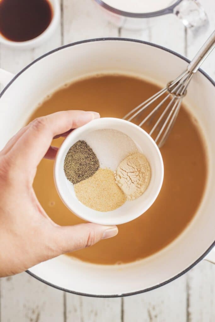 A hand holds a small bowl of four seasonings—black pepper, salt, garlic powder, and onion powder—over a pot of brown liquid with a whisk inside on a white wooden surface.