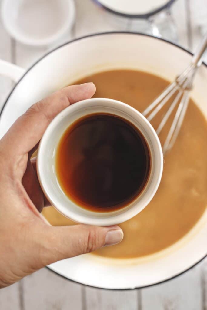A hand holds a small white cup filled with dark liquid above a bowl of light brown batter with a whisk inside.