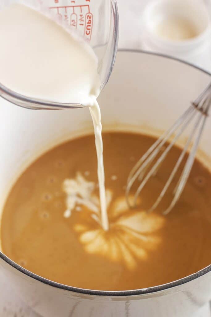 Cream is being poured from a measuring cup into a bowl of brown liquid, likely a sauce or soup, with a metal whisk resting inside the bowl.