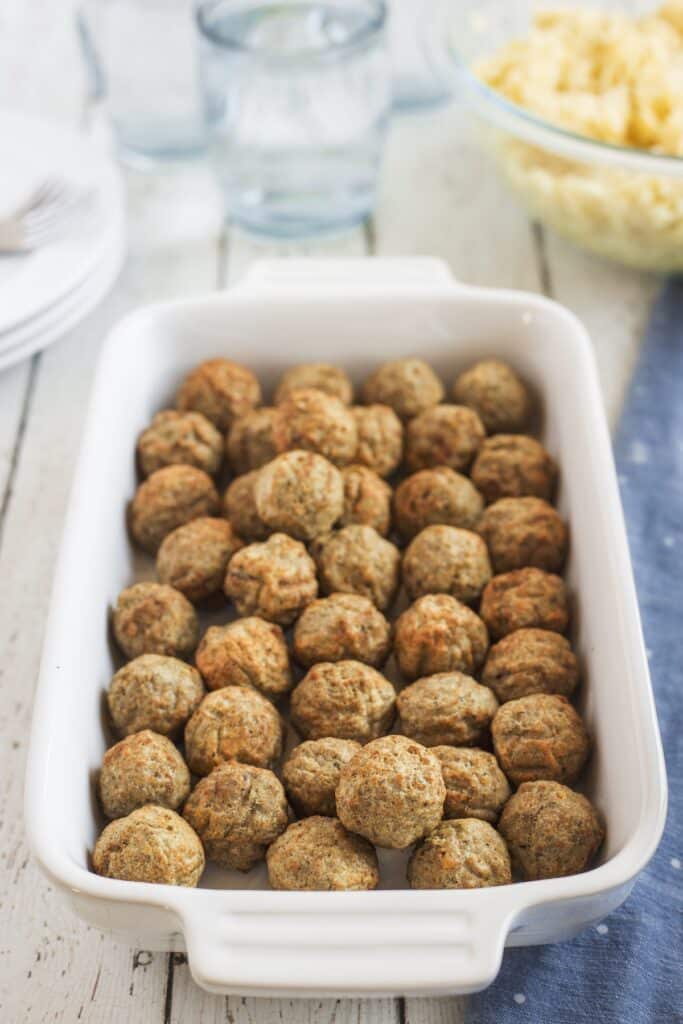 A white baking dish filled with evenly arranged cooked meatballs sits on a rustic white wooden table, with empty glass cups and a bowl of yellow food in the background.