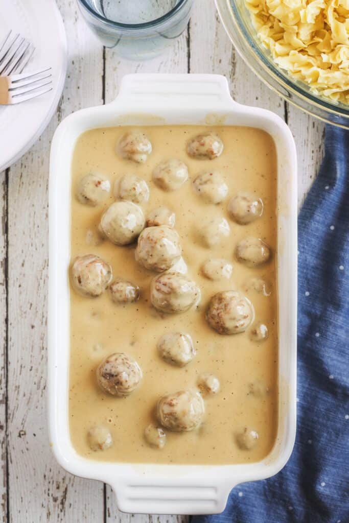 A white baking dish filled with meatballs covered in creamy beige gravy sits on a rustic white wooden table, next to a blue cloth and a bowl of uncooked egg noodles.