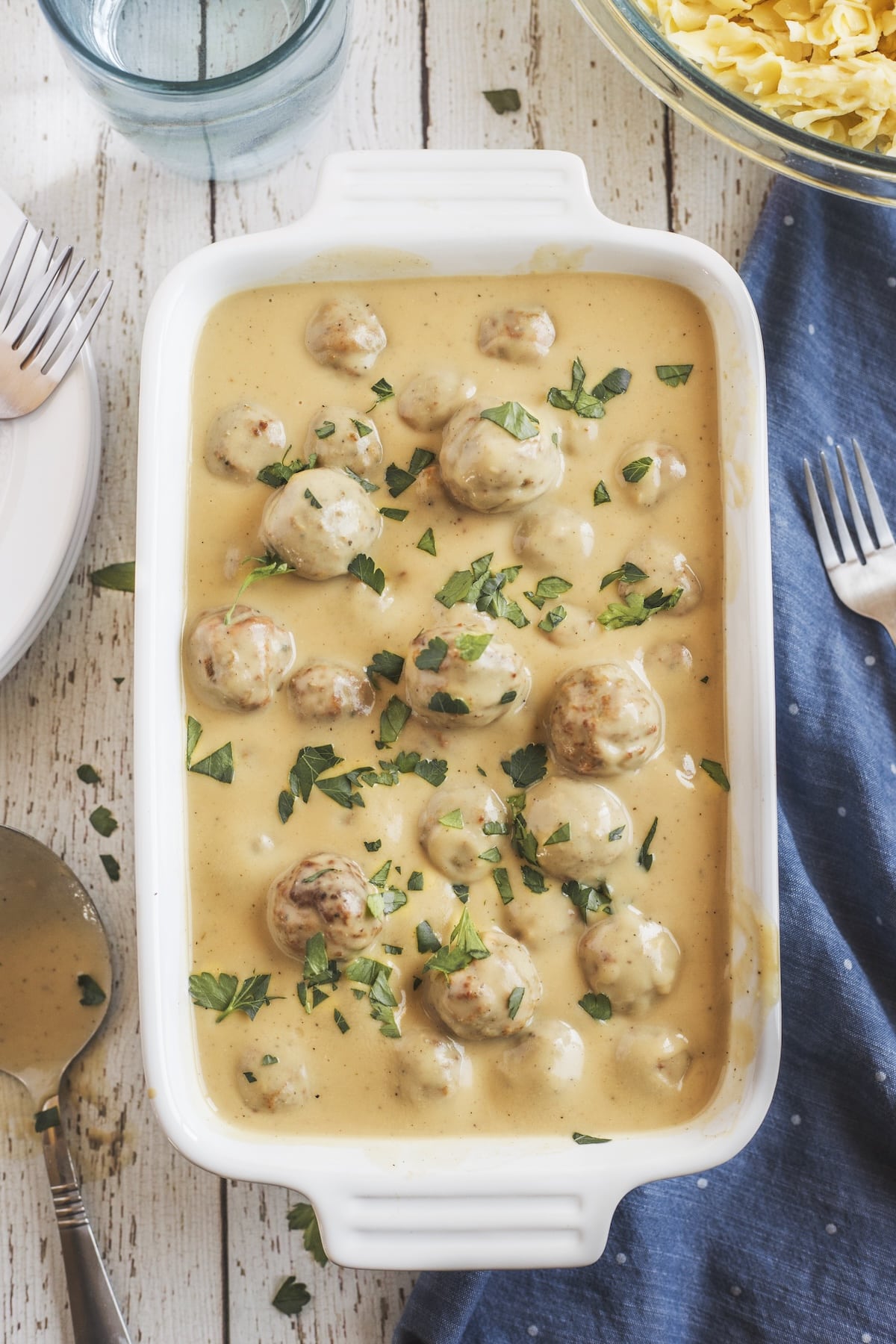 A white baking dish filled with meatballs in creamy yellow sauce, garnished with chopped parsley, sits on a wooden table beside a spoon, fork, plates, a glass of water, and a bowl of egg noodles.