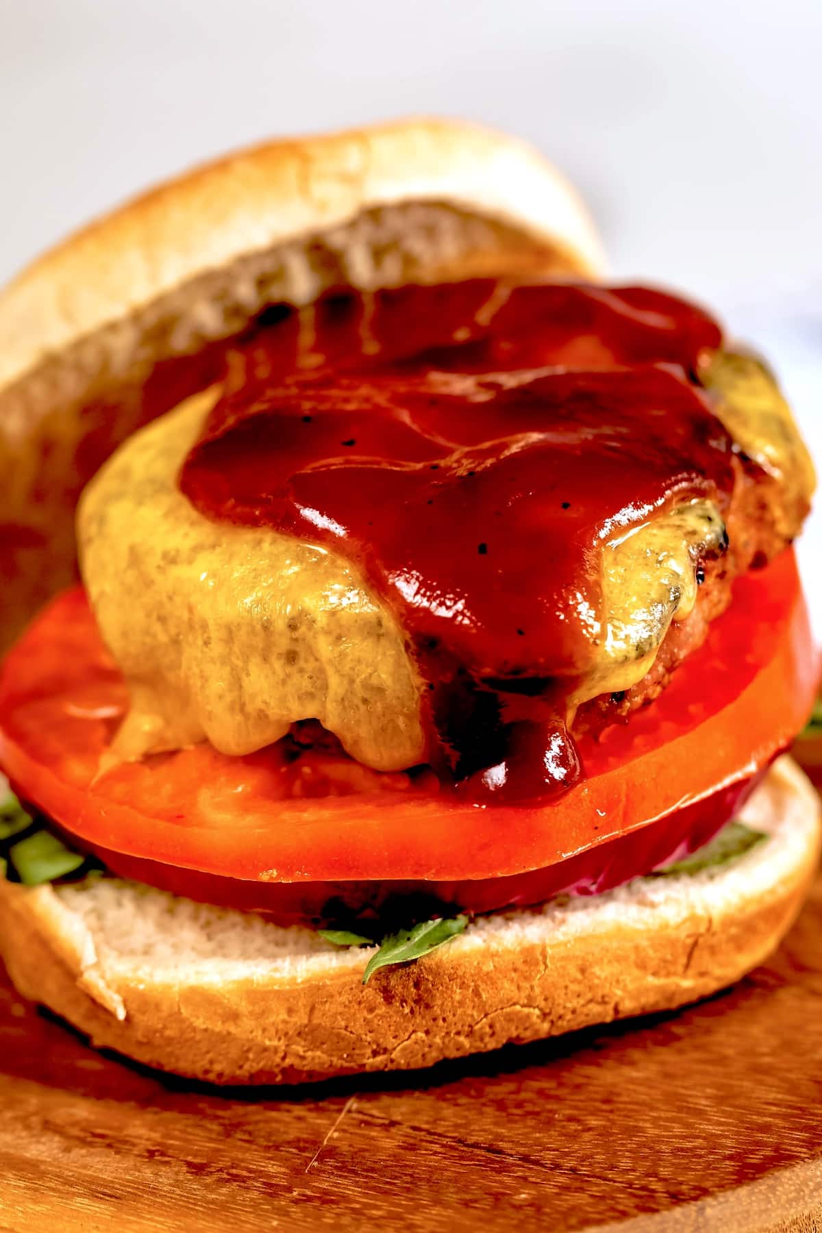 A close-up of a cheeseburger with melted cheese, barbecue sauce, fresh tomato slices, and lettuce on a sandwich bun, served on a wooden surface.