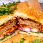 A sliced burger on a wooden board, filled with a patty, sliced red onions, tomato, cheddar cheese, and arugula, with a cup of sauce and more arugula in the background.