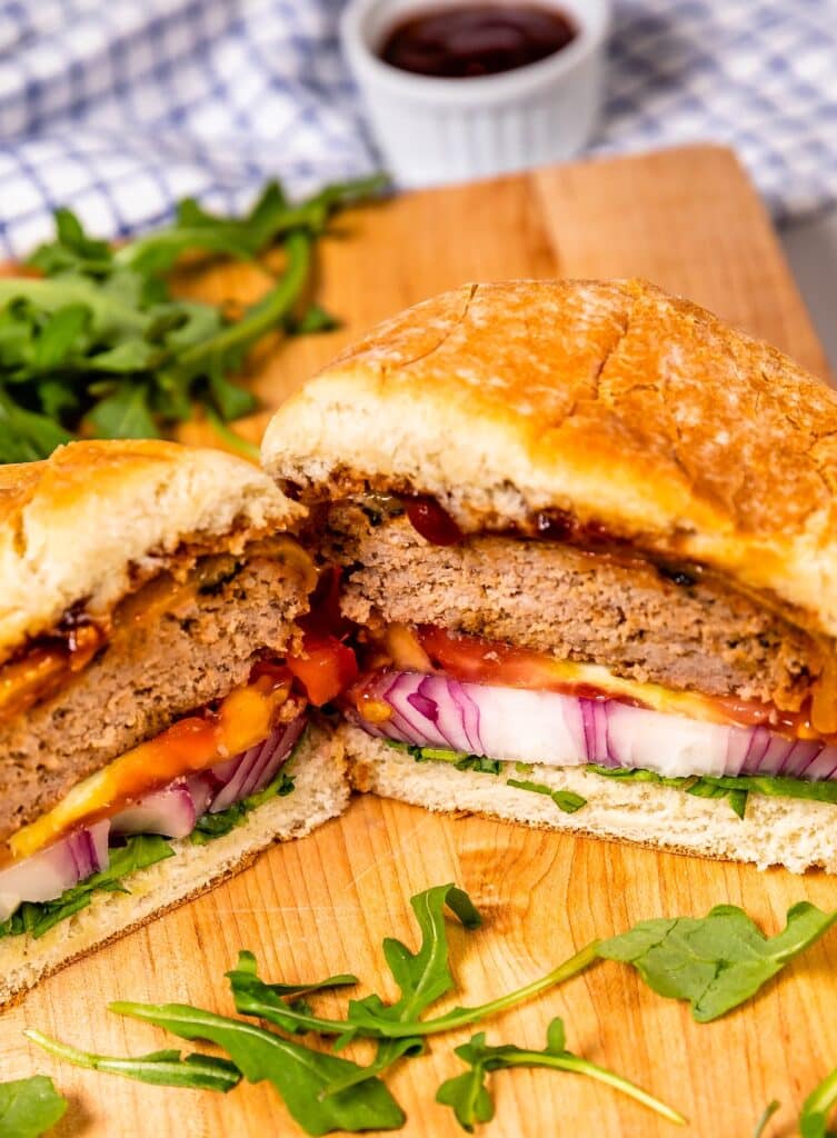 A sliced burger on a wooden board, filled with a patty, sliced red onions, tomato, cheddar cheese, and arugula, with a cup of sauce and more arugula in the background.