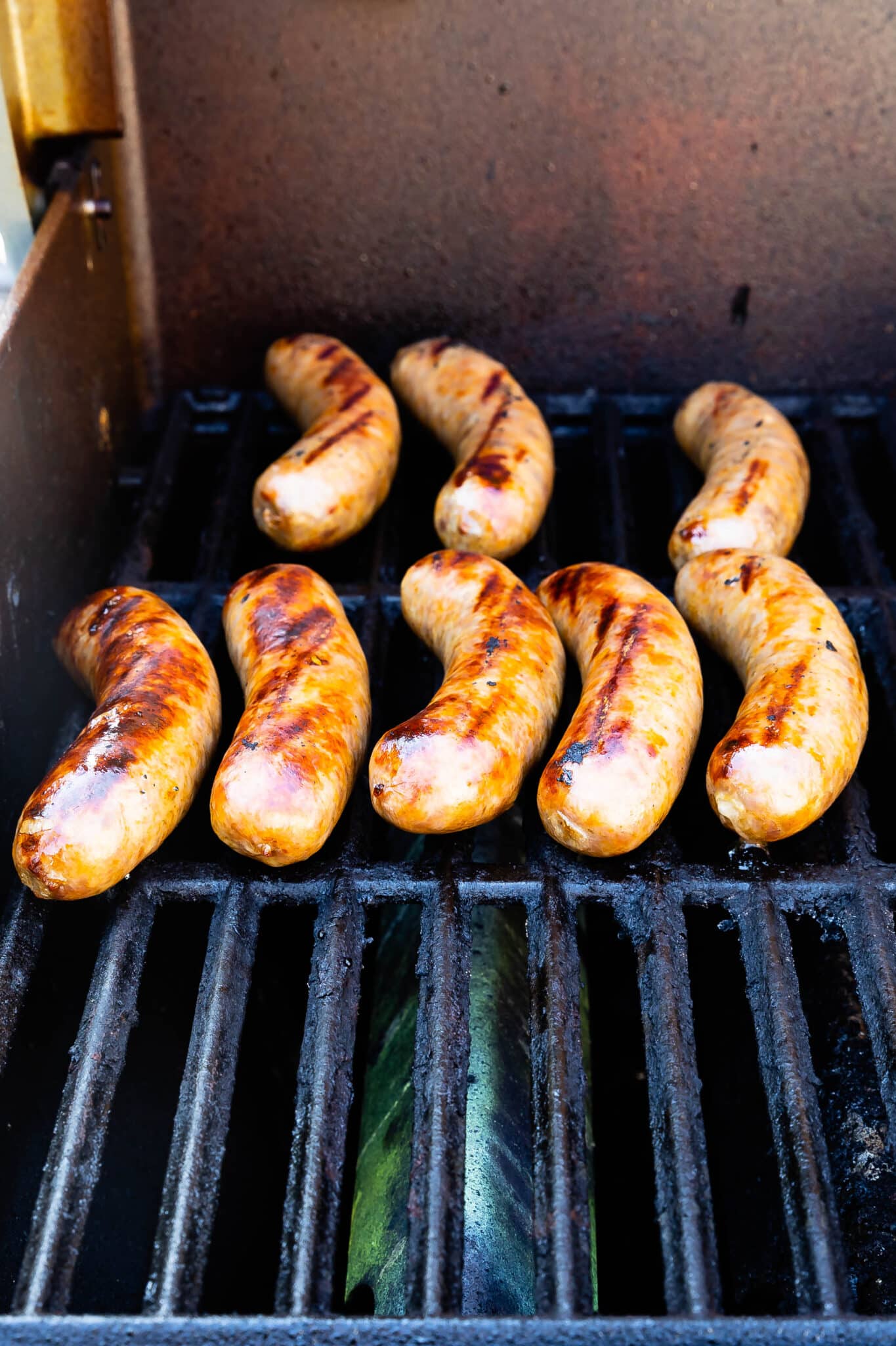 Eight sausages are being grilled on a barbecue, showing grill marks and a golden-brown, slightly charred appearance. The grill is black with visible grates and a slight flame beneath the sausages.