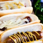 Three grilled sausages in hot dog buns are topped with mustard and sliced onions. The hot dogs are arranged closely together with a blurred background of green herbs and a blue drink can.