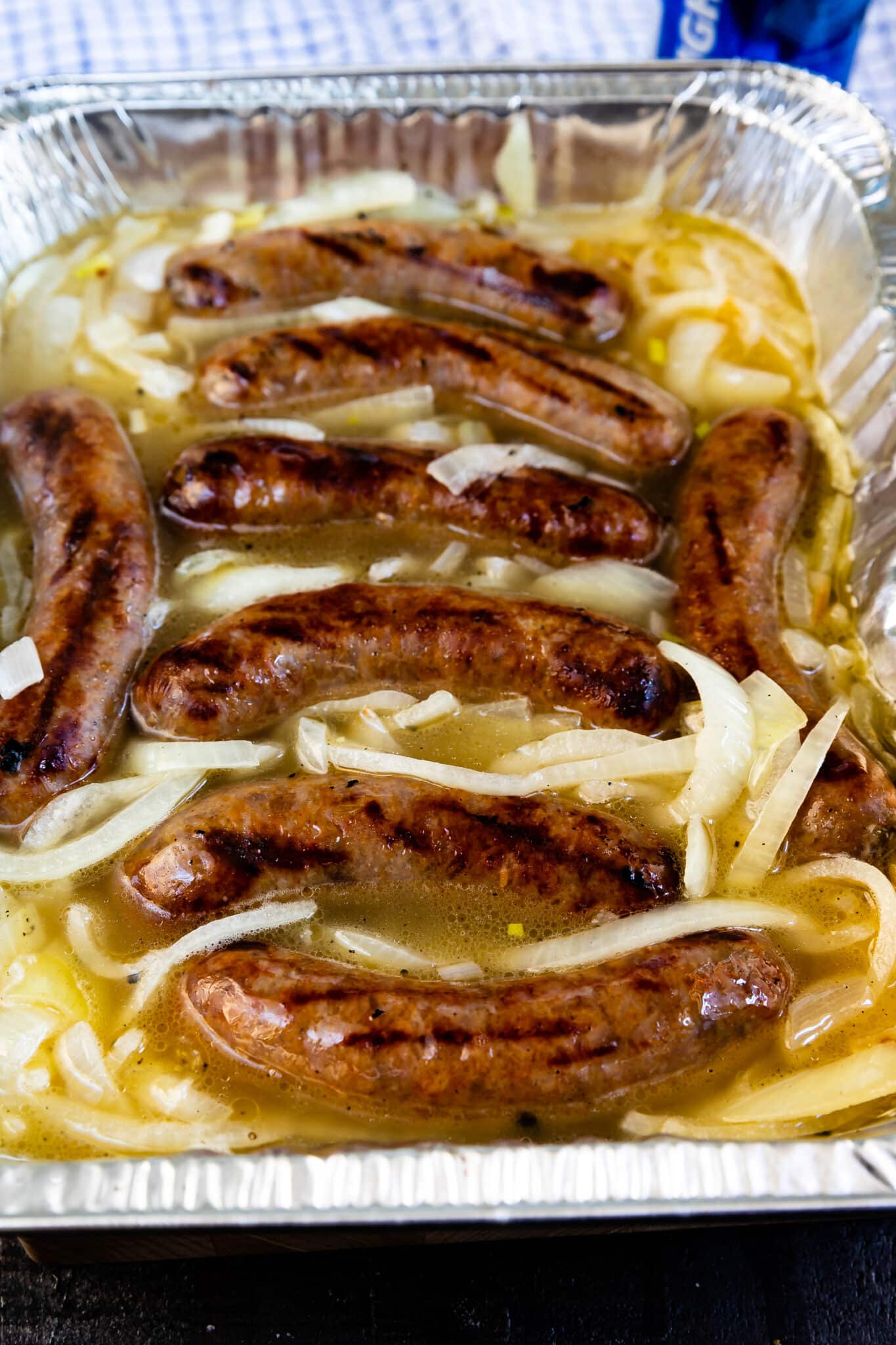 Several grilled sausages sit in a foil tray filled with sliced onions and a light broth, likely beer, ready for serving or further cooking.