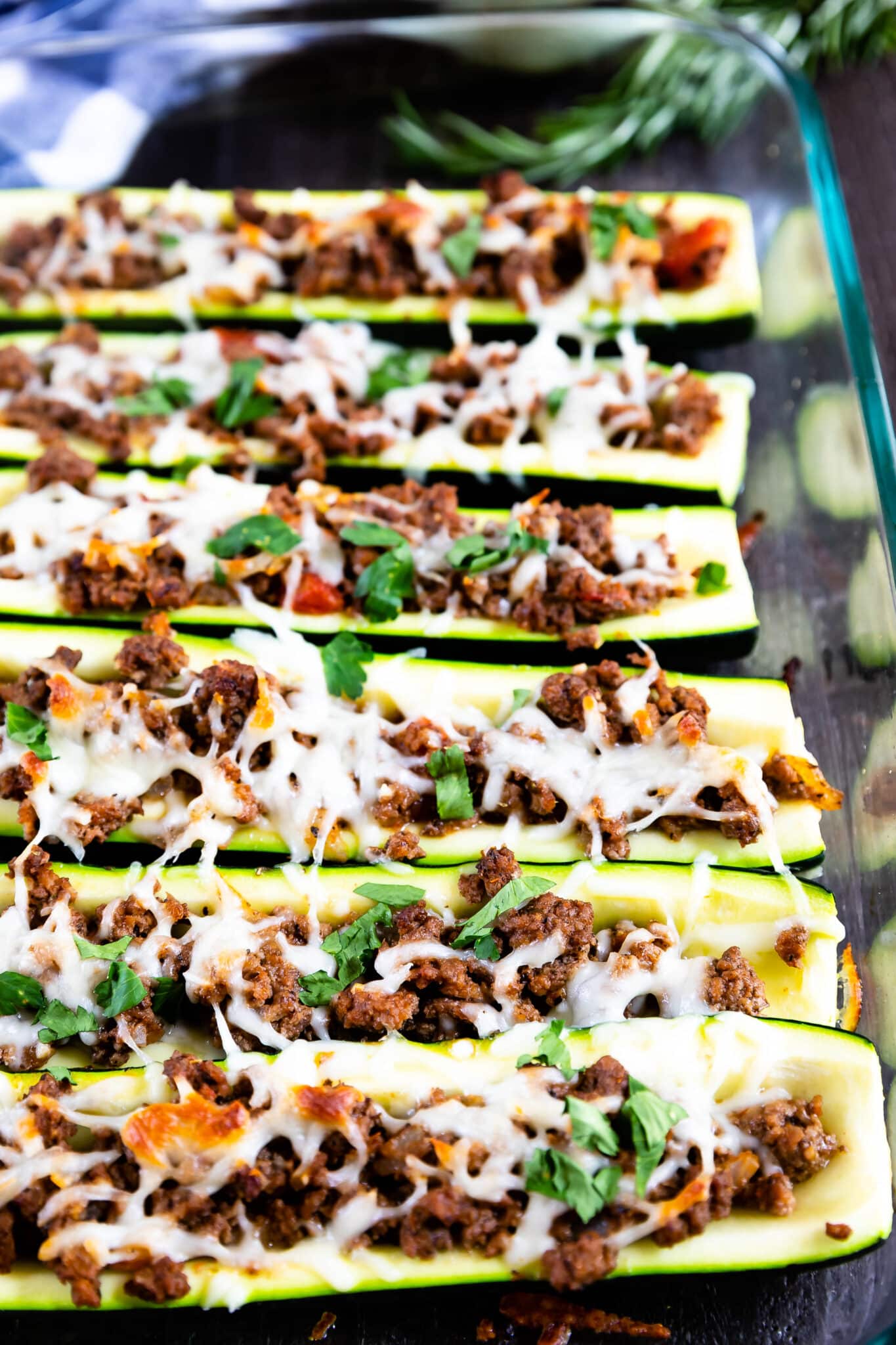 Zucchini boats filled with cooked ground meat, topped with melted cheese and chopped herbs, arranged neatly in a glass baking dish.