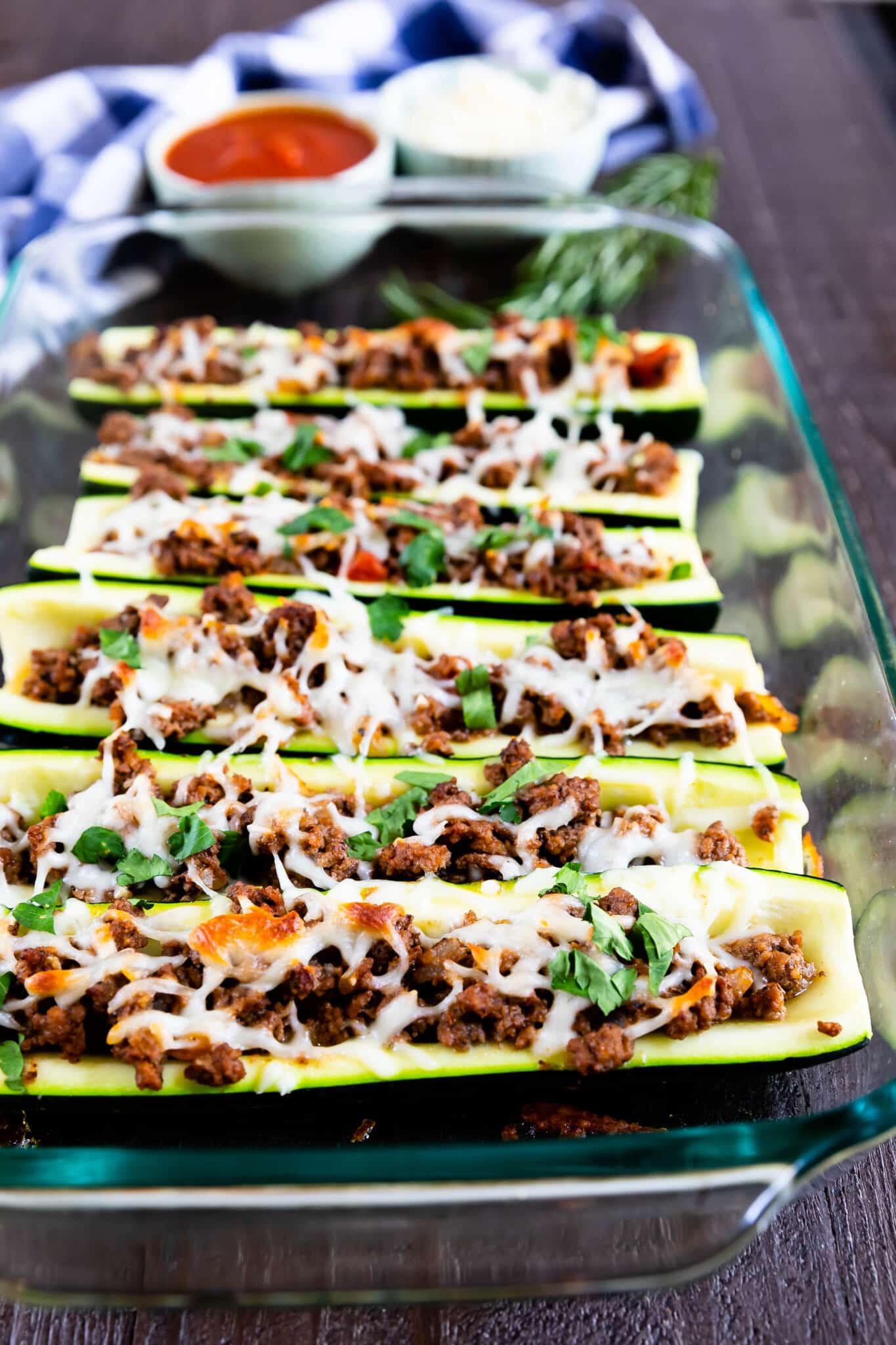 A glass baking dish filled with zucchini boats stuffed with seasoned ground meat and topped with melted cheese and herbs. In the background, there are small bowls of sauce and grated cheese.