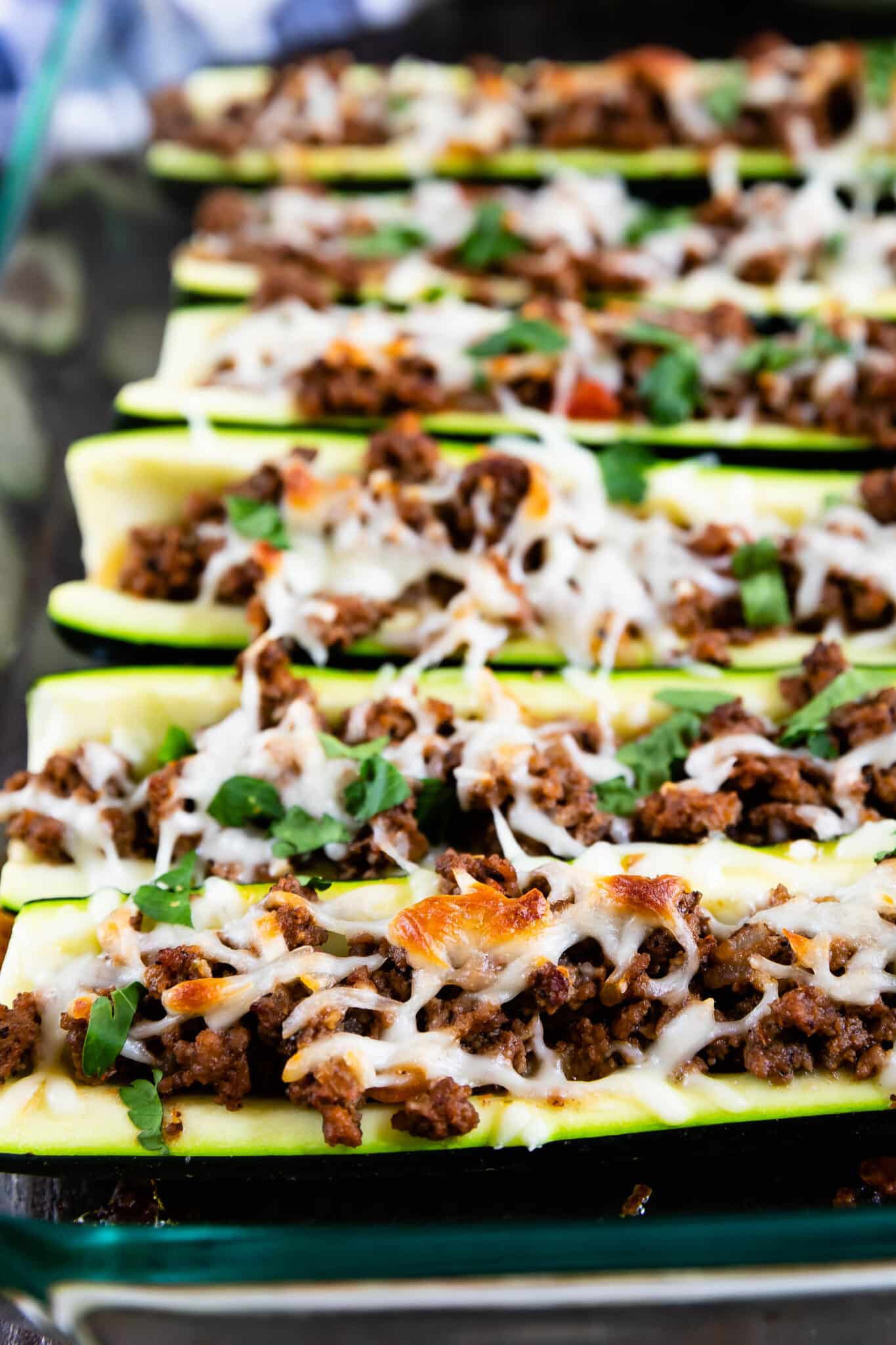 Rows of zucchini boats filled with seasoned ground beef and topped with melted cheese and chopped herbs, arranged in a glass baking dish.