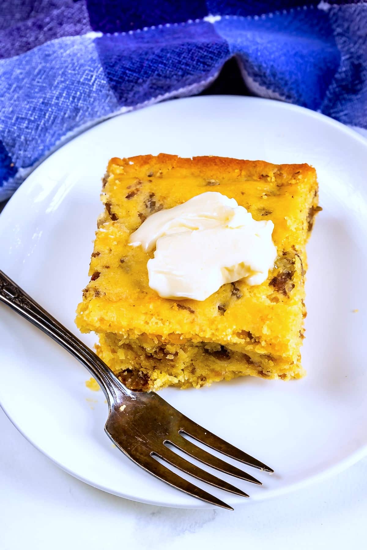A square slice of cornbread with visible bits of sausage sits on a white plate. A dollop of butter is melting on top, and a fork rests beside it. In the background, a blue and purple checkered cloth is visible.
