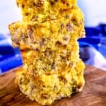 Four thick squares of sausage cornbread are stacked on a wooden cutting board, with a blurred blue and white cloth in the background. The cornbread appears moist with visible sausage pieces inside.