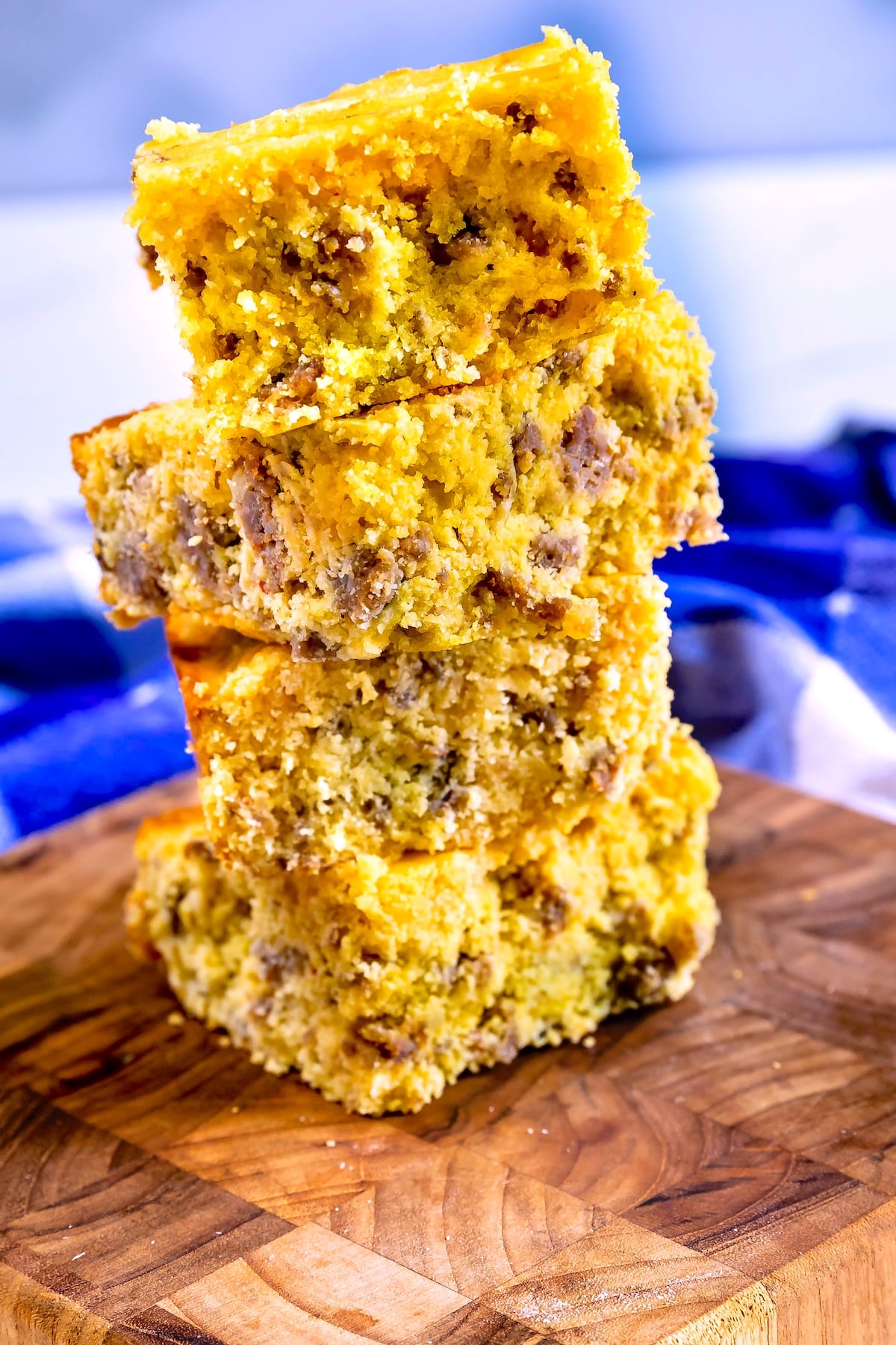 Four thick squares of sausage cornbread are stacked on a wooden cutting board, with a blurred blue and white cloth in the background. The cornbread appears moist with visible sausage pieces inside.