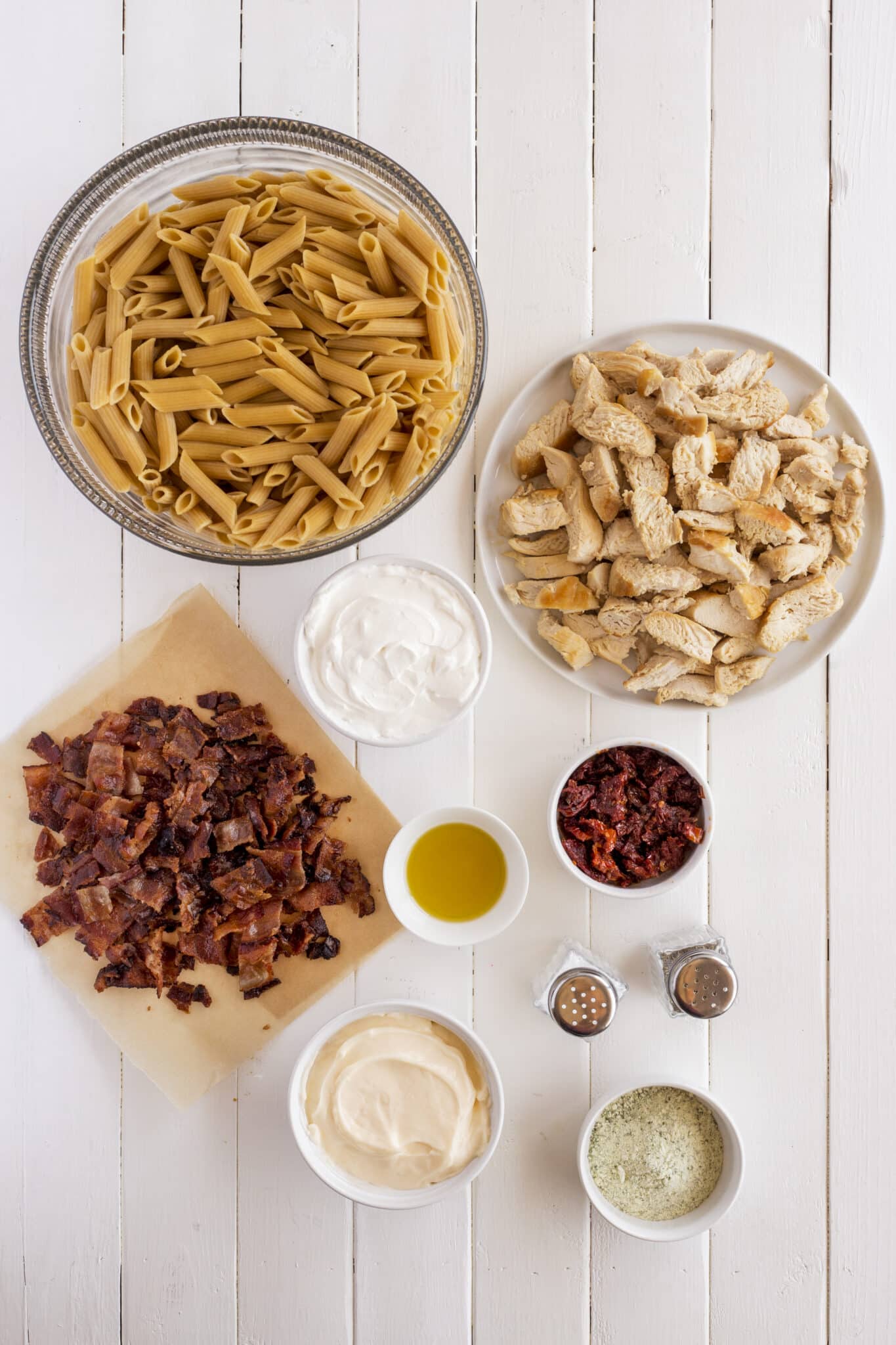 Various ingredients for a pasta dish arranged on a white wooden surface, including uncooked penne, cooked chicken pieces, chopped bacon, sun-dried tomatoes, white sauces, olive oil, ranch seasoning, and salt and pepper shakers.