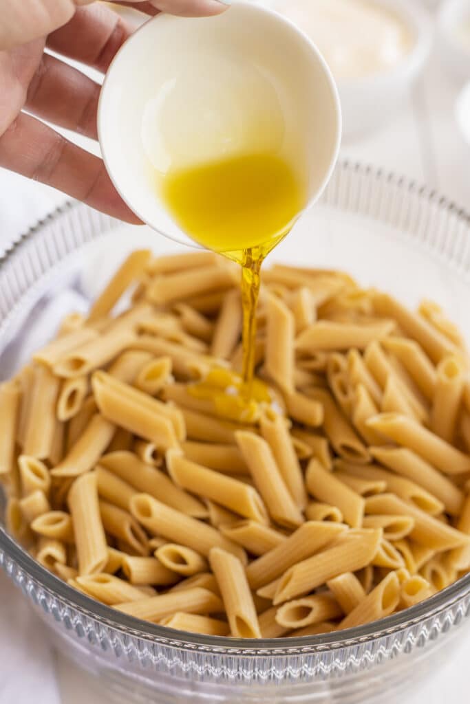 A hand pours golden olive oil from a small white bowl onto a glass bowl filled with cooked penne pasta.