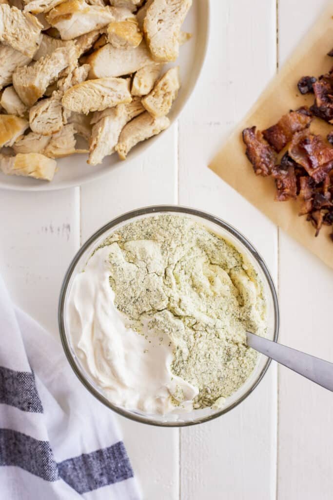A glass bowl with ranch seasoning being mixed into sour cream, next to a plate of cooked chicken pieces and a cutting board with crispy bacon on a white wooden surface.