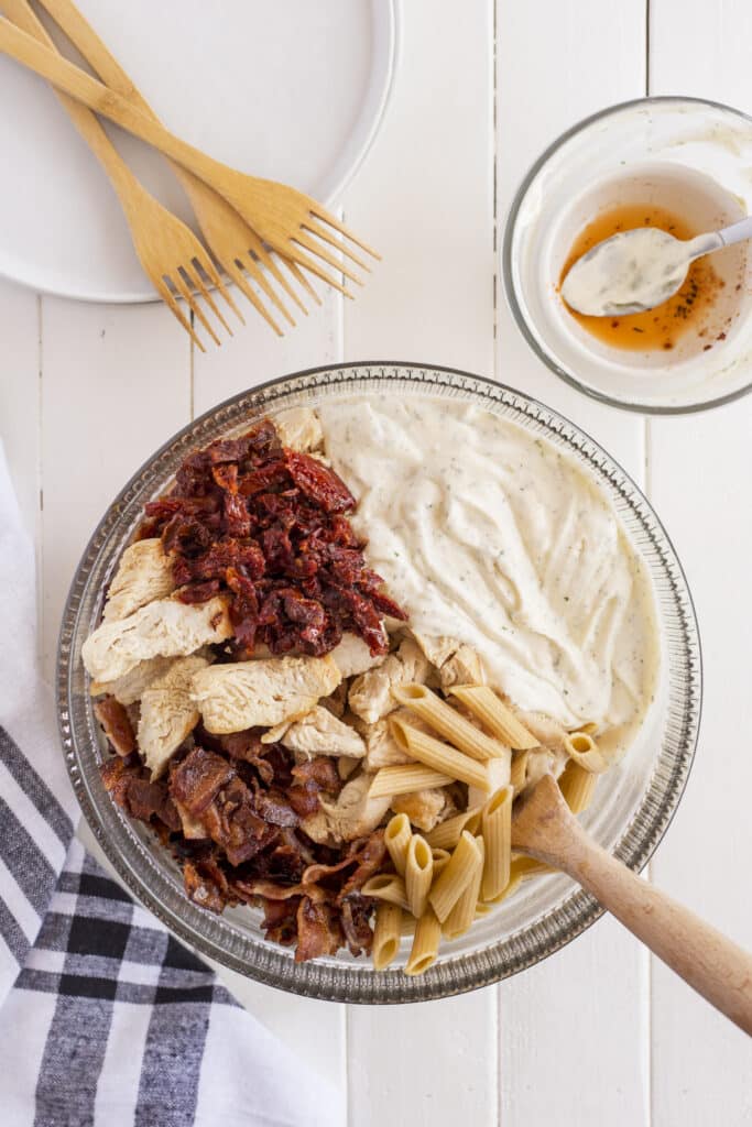 A glass bowl contains creamy white sauce, penne pasta, sliced cooked chicken, sun-dried tomatoes, and crumbled bacon, with a wooden spoon resting inside. Nearby are a striped towel, a bowl with sauce, and wooden forks on a plate.