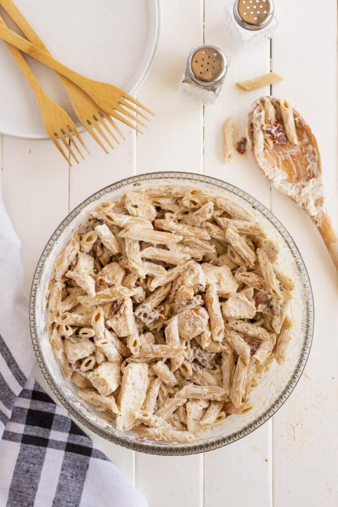 A bowl of creamy penne pasta with chicken sits on a white table, surrounded by plates, wooden forks, salt and pepper shakers, and a wooden spoon. A black-and-white checkered napkin is beside the bowl.