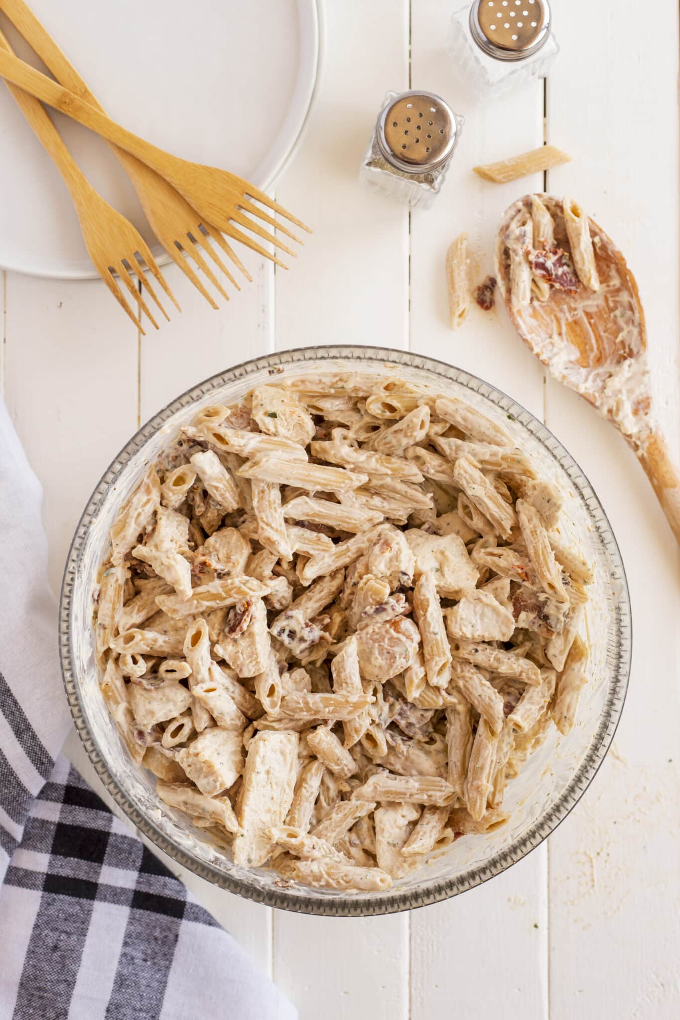 A bowl of creamy penne pasta with chicken sits on a white table, surrounded by plates, wooden forks, salt and pepper shakers, and a wooden spoon. A black-and-white checkered napkin is beside the bowl.