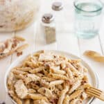 A white plate filled with creamy penne pasta with pieces of chicken and sun-dried tomatoes sits on a white table. In the background are a bowl of more pasta, salt and pepper shakers, two empty glasses, and wooden forks.