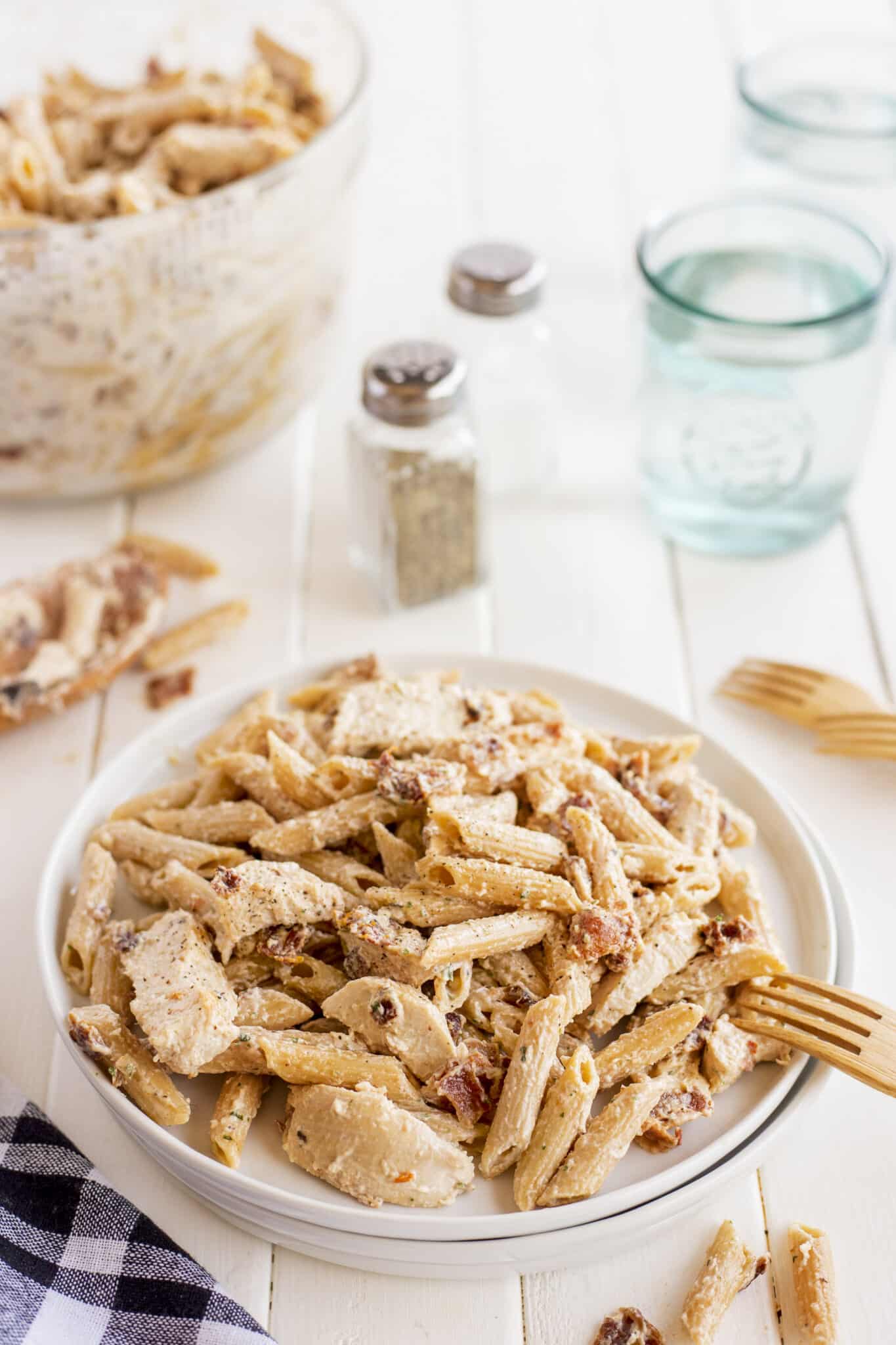 A white plate filled with creamy penne pasta with pieces of chicken and sun-dried tomatoes sits on a white table. In the background are a bowl of more pasta, salt and pepper shakers, two empty glasses, and wooden forks.