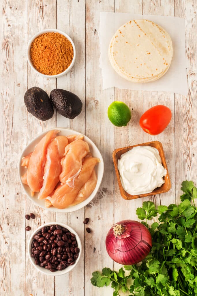 Overhead view of ingredients on a wooden surface: raw chicken strips, black beans, taco seasoning, avocados, lime, tomato, flour tortillas, sour cream, red onion, and fresh cilantro.