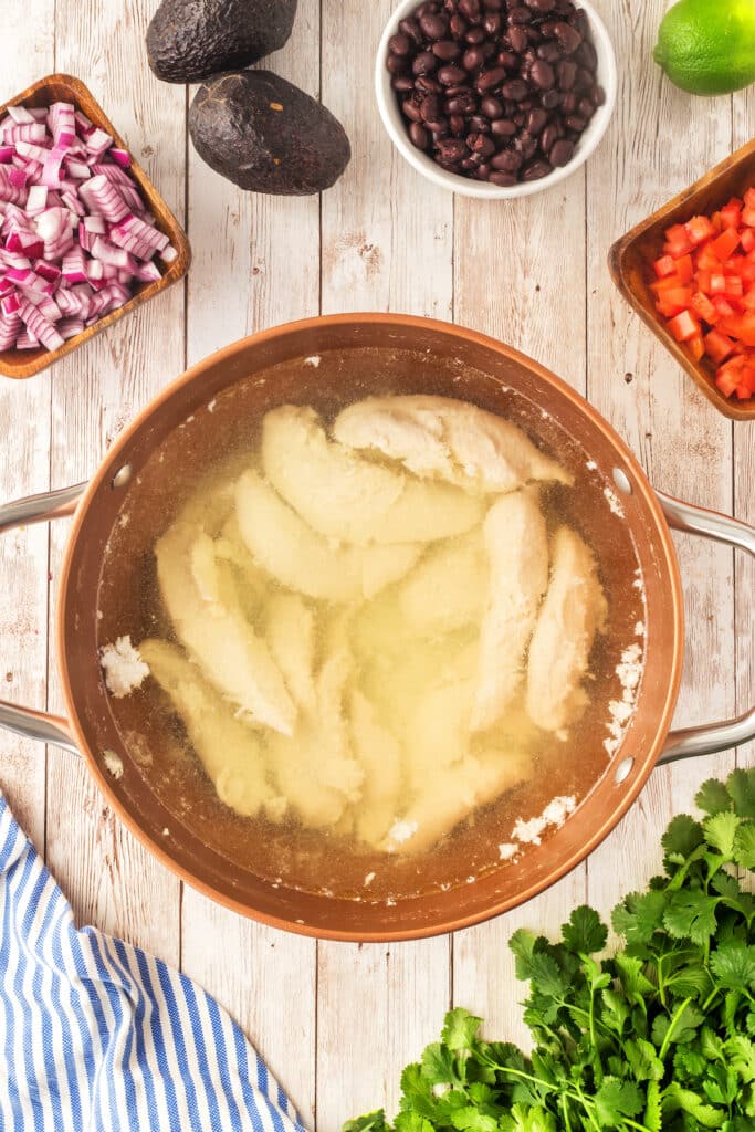 A pot of raw chicken breasts boiling in water sits on a wooden table, surrounded by chopped red onion, black beans, diced red bell pepper, cilantro, avocado, a lime, and a blue-striped cloth.